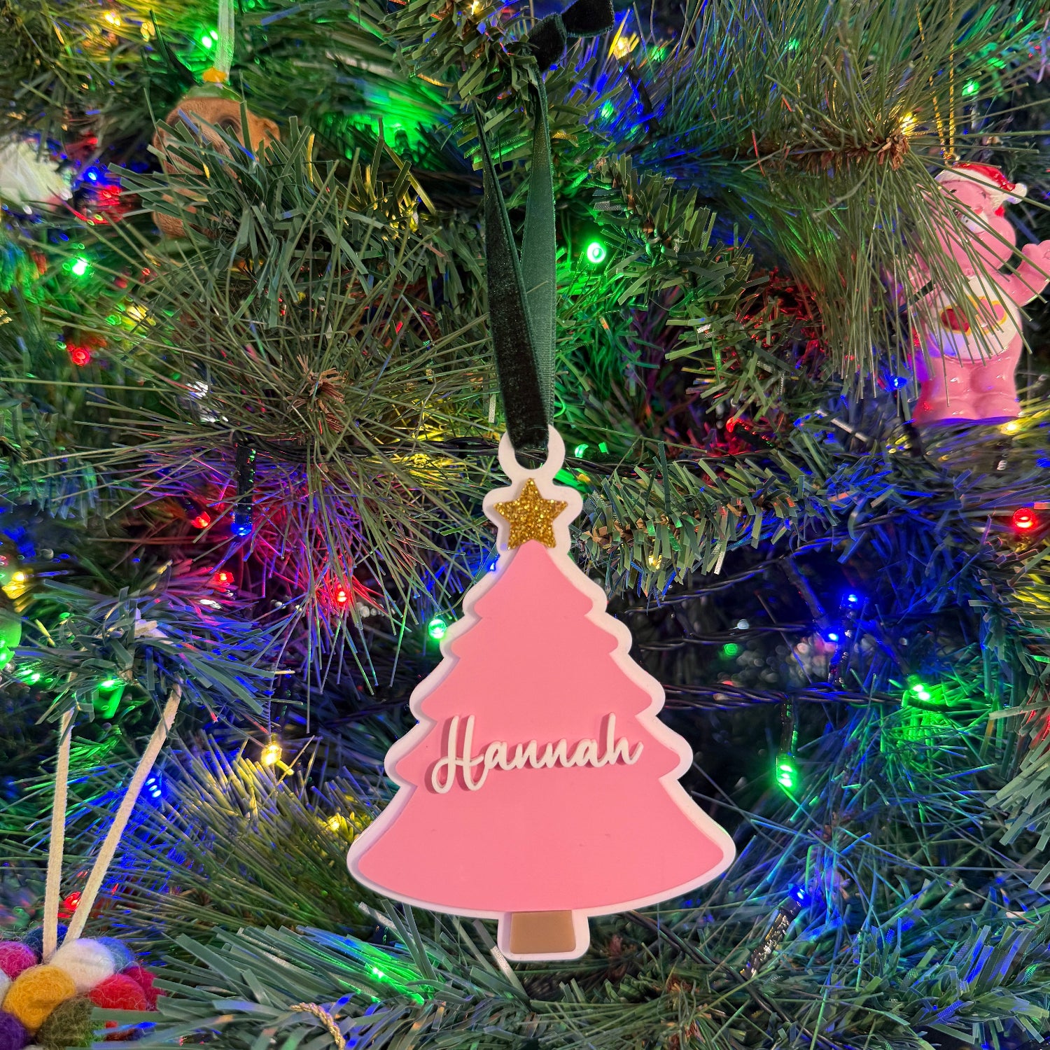 Decorative pink Christmas tree ornament with 'Hannah' on a lit Christmas tree.