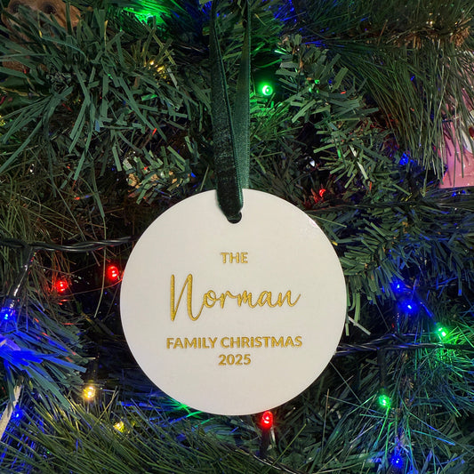 Round white ornament with gold text "The Norman Family Christmas 2025" on a Christmas tree with lights.