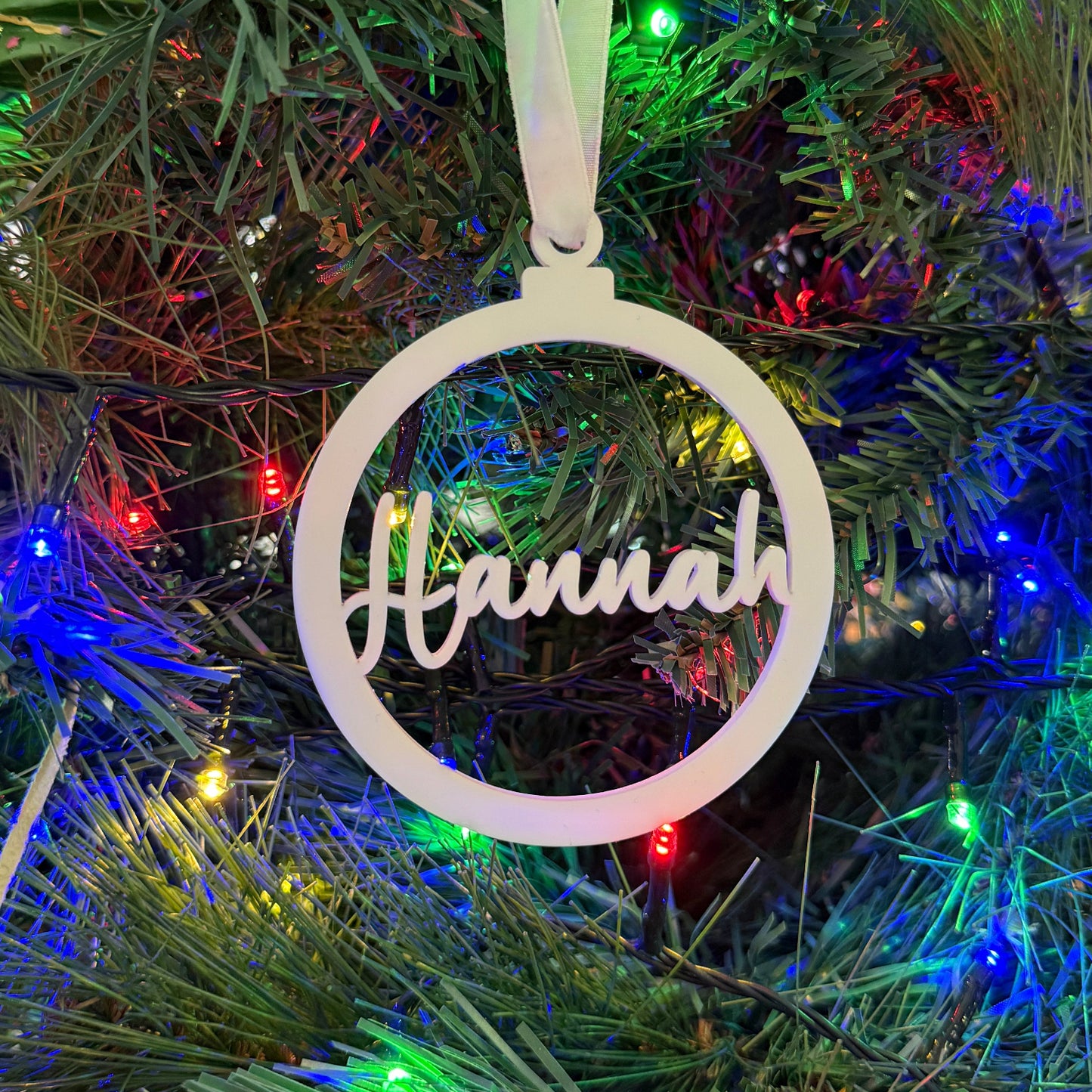 White circular ornament with 'Hannah' on a Christmas tree with colorful lights.